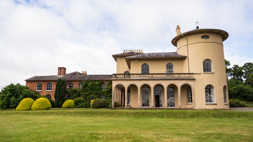 View of Cronkhill, a cream-coloured Italianate villa with a distinctive circular tower, arched loggia and balustraded terrace, seen across a wide lawn with clipped shrubs and mature trees.